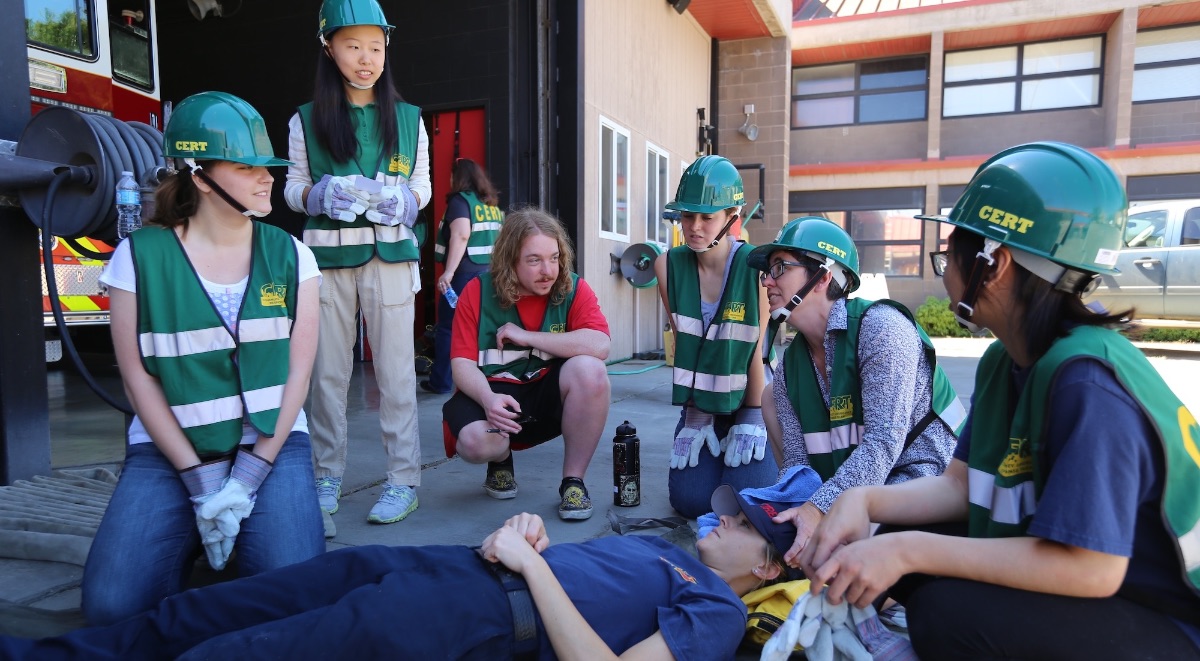 A group of people wearing matching safety vests and hard hats gather around a person laying on the ground outside of the UC Davis Fire building